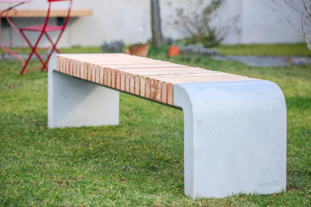 Moderne Sitzbank mit abgerundeten Füßen aus Beton und längs verlaufenden Holzlatten steht in einer öffentlichen Grünanlage.