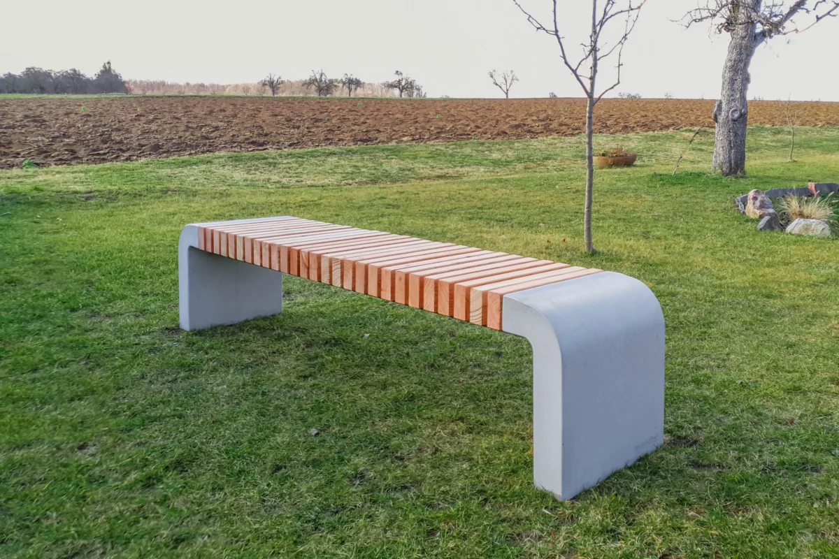 Moderne Sitzbank mit abgerundeten Füßen aus Beton und längs verlaufenden Holzlatten steht in einer öffentlichen Grünanlage.