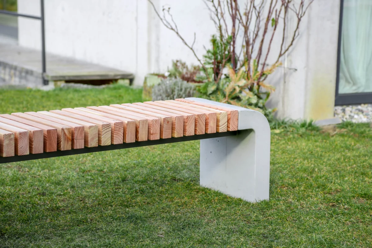 Moderne Sitzbank mit abgerundeten Füßen aus Beton und längs verlaufenden Holzlatten steht in einer öffentlichen Grünanlage.
