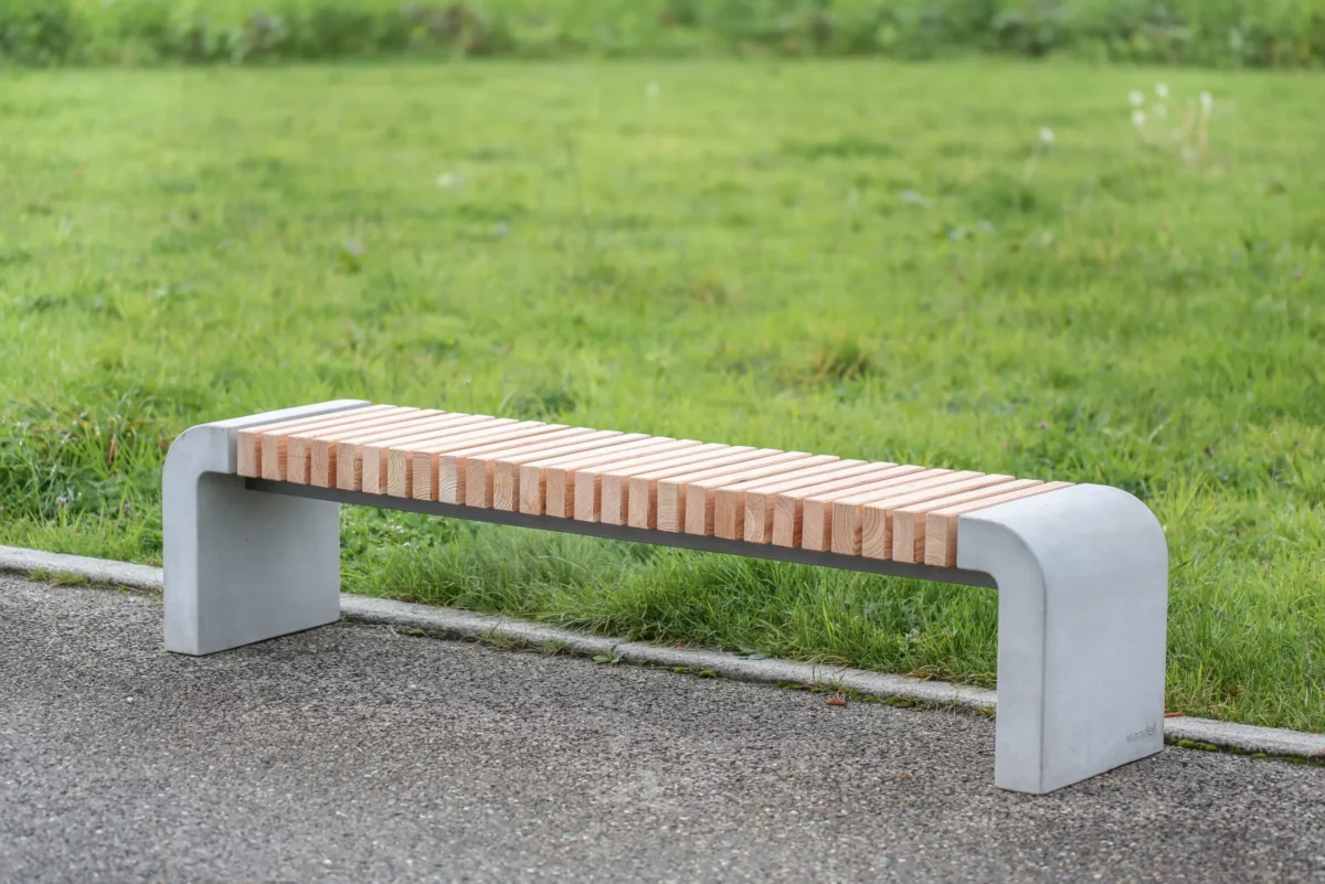 Moderne Sitzbank mit abgerundeten Füßen aus Beton und längs verlaufenden Holzlatten steht in einer öffentlichen Grünanlage.