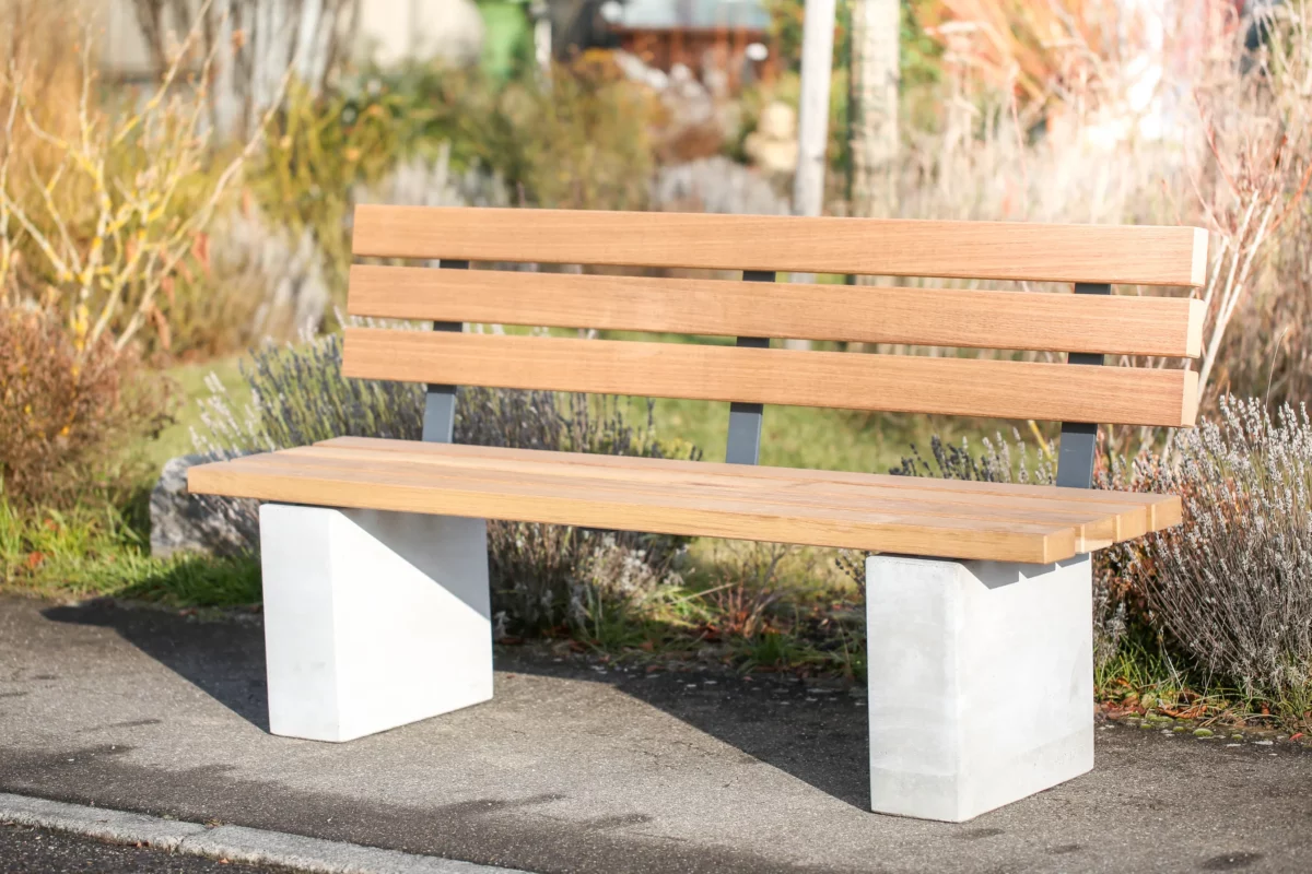 Rollstuhlgerechte Sitzbank aus Hartholz und nach innen versetzten Standfüßen aus Beton steht auf einer öffentlichen Fläche mit Begrünung im Hintergrund