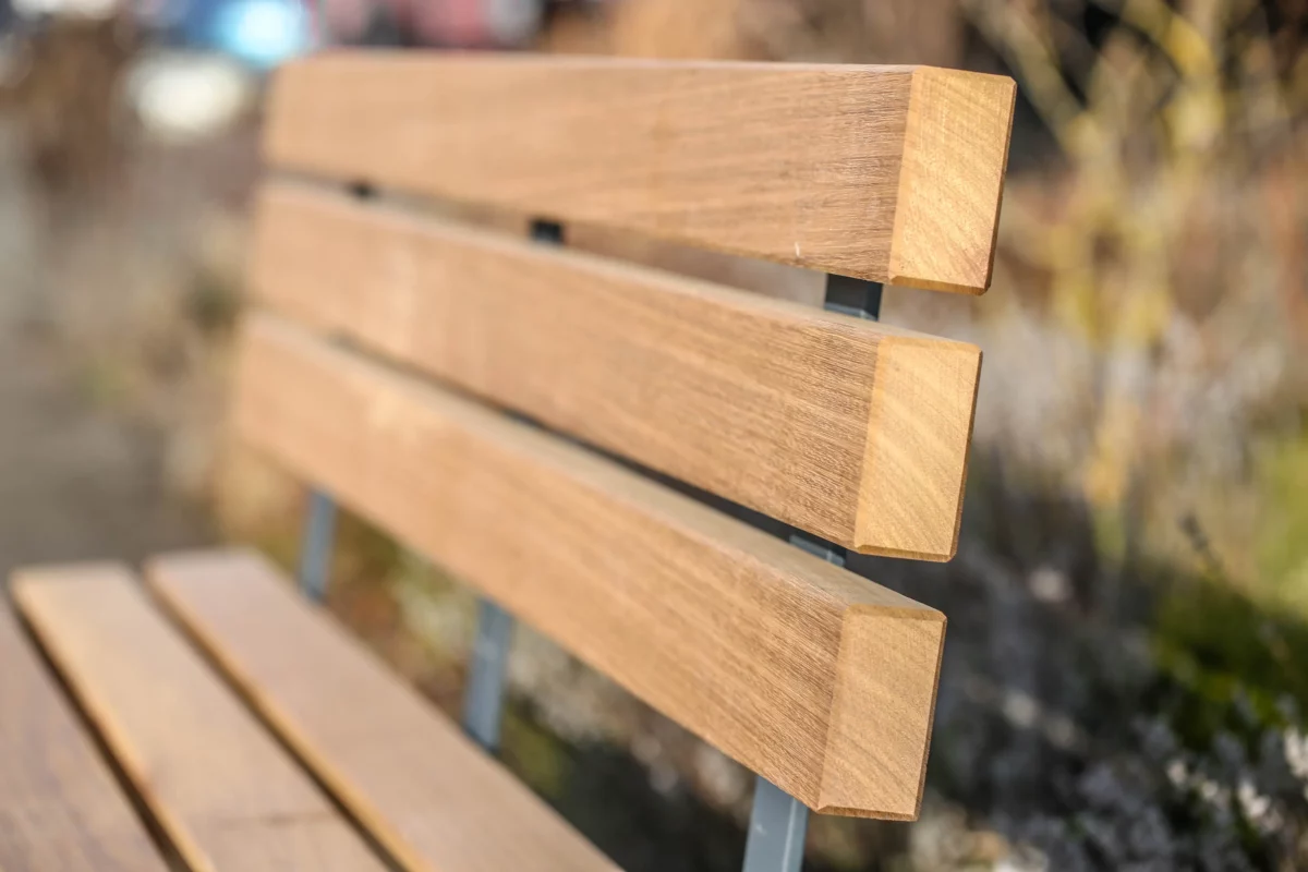 Rollstuhlgerechte Sitzbank aus Hartholz und nach innen versetzten Standfüßen aus Beton steht auf einer öffentlichen Fläche mit Begrünung im Hintergrund